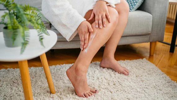 Person cremt Bein ein, sitzt auf einem Sofa im Wohnzimmer, neben einem Tisch mit Pflanze.